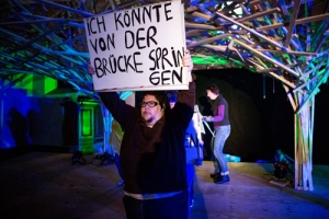Ein Mann steht auf einer Bühne und hält ein Schild hoch. Auf dem Schild steht: "Ich könnte von der Brücke springen".