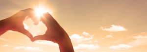 Zwei Hände sind zum Herz geformt. Im Hintergrund sieht man die Sonne.