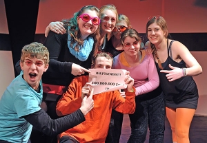Jugendliche stehen im Pulk zusammen. Sie tragen rosa Sonnenbrillen und halten ein Schild hoch. Auf dem Schild steht: "Hauptgewinn!!! 100.000.000 Euro!!!"