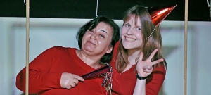 Zwei Frauen lehnen sich aneinander und lächeln in die Kamera. Sie tragen rote Kleidung. Die rechte Frau trägt ein rotes Geburtstagshütchen und macht das "Peace"-Zeichen.
