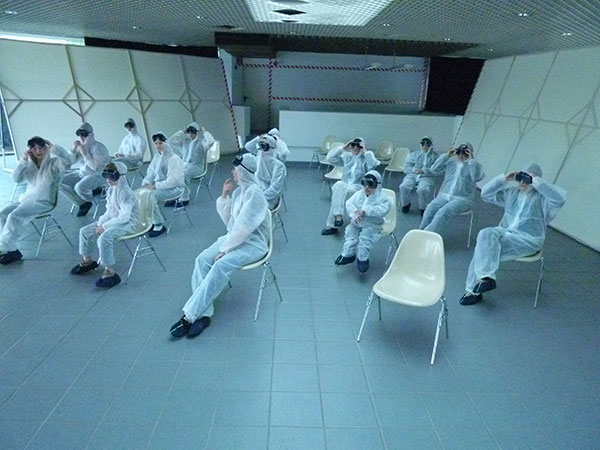 Filmrojekte-und-Kurzfilme-(20) Personen sitzen in Laborkitteln und mit schwarzen Taucherbrillen auf Stühlen in einem leeren Raum.