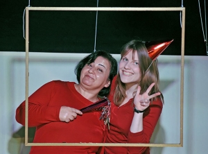 Zwei Frauen lehnen sich aneinander und lächeln in die Kamera. Sie tragen rote Kleidung. Die rechte Frau trägt ein rotes Geburtstagshütchen und macht das "Peace"-Zeichen.