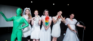 Frauen in Hochzeitskleidern stehen nebeneinander auf einer Bühne und lächeln. Sie halten Putz-Utensilien, wie einen Staubwedel und einen Staubsauger in der Hand. Vor ihnen steht eine Person in einem grünen Ganzkörper-Suit.