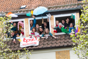 Viele verkleidete Menschen winken aus einem Fenster.