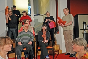 Viele Personen sitzen und stehen zusammen im Raum. Ein Mann lehnt schlafend an der Wand. Zwei Frauen sitzen sich im Schneidersitz gegenüber. Ein Mann häkelt im Stehen.