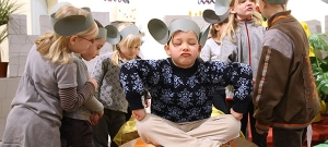 Ein Kind sitzt im Schneidersitz auf einem Tisch und schließt trotzig die Augen. Um das Kind herum stehen weitere Kinder. Alle tragen selbstgebastelte Mäuseohren auf dem Kopf.