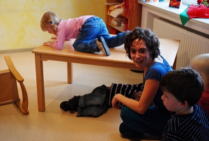 Eine Frau sitzt mit zwei Kindern in einem Raum. Eines der Kinder krabbelt über einen Tisch.