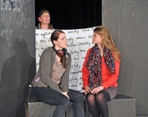 Zwei Frauen sitzen nebeneinander und schauen sich an. Hinter ihnen steht eine Person und hält ein Banner in die Luft. Auf dem Banner steht ein englischer Text.
