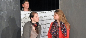 Zwei Frauen sitzen nebeneinander und schauen sich an. Hinter ihnen steht eine Person und hält ein Banner in die Luft. Auf dem Banner steht ein englischer Text.