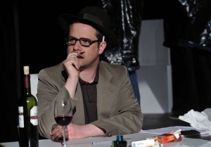 Ein Mann sitzt mit einem Glas Wein am Schreibtisch. Er hält einen Stift in der Hand und nimmt eine nachdenkliche Pose ein. Hinter seinem Ohr klemmt eine Zigarette.