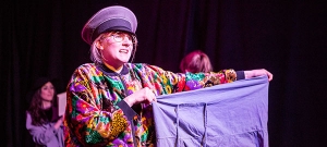 Eine junge Frau mit Schirmmütze und bunter Jacke hält ein lila Kleidungsstück vor sich ausgebreitet wie ein rotes Tuch beim Torero.