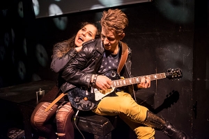 Ein Mann mit Lederjacke spielt auf einer E-Gitarre. Eine Frau lehnt sich an seinem Rücken an.
