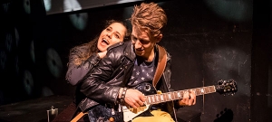 Ein Mann mit Lederjacke spielt auf einer E-Gitarre. Eine Frau lehnt sich an seinem Rücken an.