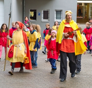 Kinder in Kostümen folgen zwei Stadtführer*innen. Die Stadtführer*innen sind ebenfalls kostümiert. Einer der Stadtführer*innen spielt Okulele.