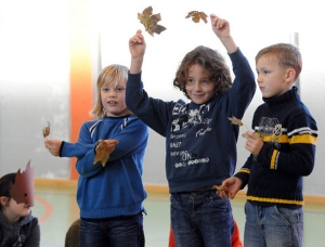 Drei Kinder stehen nebeneinander und halten Laubblätter in ihren Händen. Neben ihnen hockt ein weiteres Kind mit einer selbstgebastelten Blätter-Maske vor dem Gesicht.
