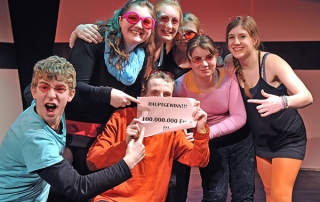 Jugendliche stehen im Pulk zusammen. Sie tragen rosa Sonnenbrillen und halten ein Schild hoch. Auf dem Schild steht: "Hauptgewinn!!! 100.000.000 Euro!!!"