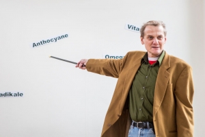 Ein Mann in braunem Jackett zeigt mit einem Stock auf Wörter an der Wand. An der Wand steht: "Anthocyane", "Vitamine" und "Freie Radikale".