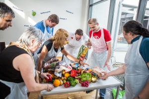 Viele Personen stehen um einen Tisch herum und sortieren Essen, das auf dem Tisch liegt. Die Personen tragen weiße Schutzanzüge.