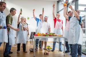 Viele Personen stehen um einen Tisch herum und halten Obst und Gemüse in die Luft. Auf dem Tisch liegt ein großer Haufen mit weiterem Obst und Gemüse. Die Personen tragen weiße Schutzkittel.