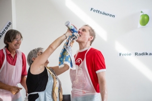 Eine Frau sprüht einem Mann Sahne in den Mund. Ein weiterer Mann sieht lachend zu.