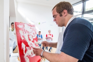 Ein Mann trägt mit einem Spachtel eine weiße Masse auf eine rote Leinwand auf.