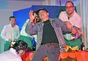 Ein Mann lässt sich lachend auf einen Esstisch fallen. Mehrere Personen stehen um ihn herum und halten ihn fest.