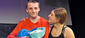 Eine Frau und ein Mann stehen lächelnd zusammen auf der Bühne. Der Mann hält Decken wie ein Baby vor sich. Die Frau legt dem Mann die Hand auf die Schulter.