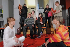 Viele Personen sitzen und stehen zusammen im Raum. Ein Mann lehnt schlafend an der Wand. Zwei Frauen sitzen sich im Schneidersitz gegenüber. Ein Mann häkelt im Stehen.