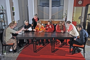 Viele Menschen sitzen zusammen an einem Esstisch. Eine Frau deckt den Tisch.