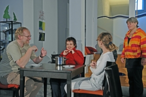 Drei Personen sitzen zusammen am Esstisch. Eine weitere Frau kommt dazu.