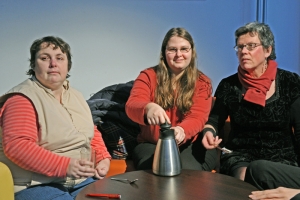 Drei Personen sitzen zusammen an einem Kaffeetisch. Die Frau in der Mitte öffnet eine Kaffeekanne.