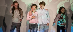 Zwei Kinder stehen Arm in Arm auf einer Bühne. Links und rechts von ihnen stehen weitere Kinder.