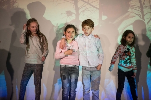 Zwei Kinder stehen Arm in Arm auf einer Bühne. Links und rechts von ihnen stehen weitere Kinder.