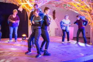 Zwei Frauen stehen Rücken an Rücken aneinander. Sie tragen große, blaue Plastiktüten auf dem Rücken. Um sie herum stehen weitere Personen.
