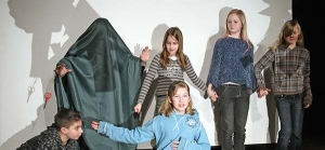 Mehrere Kinder stehen auf der Bühne und halten sich an den Händen. Eines der Kinder versteckt sich unter einer grünen Decke.