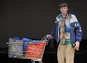 Ein Mann mit einem Einkaufswagen steht auf einer Bühne. Im Einkaufswagen stehen mehrere Tüten. Der Mann hält eine Bierflasche in der Hand. Er trägt Kleidung, die nicht zueinander passt.