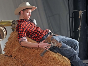 Ein Mann liegt auf einem Strohballen. Der Mann ist als Cowboy verkleidet. Er spielt mit einer Pistole, die an seinem Gürtel hängt.