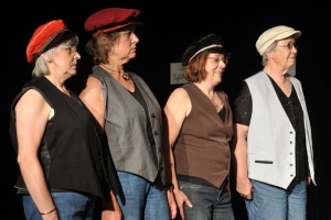 Vier Frauen stehen in einer Reihe und blicken in die selbe Richtung. Sie tragen Westen und Schirmmützen. Der Hintergrund ist schwarz.