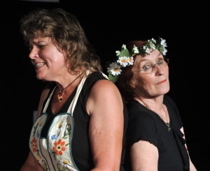 Zwei Frauen stehen Rücken an Rücken. Die linke Frau hat einen schmerzverzerrten Gesichtsausdruck. Sie trägt eine Kochschürze mit Blumen-Stickereien. Die rechte Frau schaut in die Kamera. Sie trägt einen Blumenkranz auf ihrem Kopf. Der Hintergrund ist schwarz.