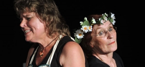 Zwei Frauen stehen Rücken an Rücken. Die linke Frau hat einen schmerzverzerrten Gesichtsausdruck. Sie trägt eine Kochschürze mit Blumen-Stickereien. Die rechte Frau schaut in die Kamera. Sie trägt einen Blumenkranz auf ihrem Kopf. Der Hintergrund ist schwarz.
