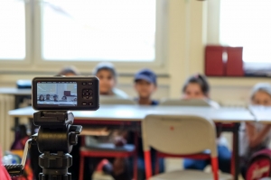 Mehrere Kinder schauen hinter einem Schultisch hervor. Sie sind nur verschwommen zu erkennen. Im Vordergrund des Bildes steht eine Kamera, die die Kinder aufnimmt.