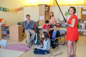 Kinder und Erwachsene sitzen in einem Klassenraum und drehen einen Film. In ihrer Mitte steht eine Kamera. Rechts im Bild steht eine Frau mit Kopfhörern und einer Tonangel in der Hand.