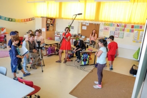 Das Foto wurde in einem Klassenzimmer aufgenommen. Eine Frau mit Kopfhörern hält eine Tonangel. Vor ihr steht eine Kamera. Kinder und ein Mann sitzen und stehen im Kreis um die Kamera herum. Ein Junge hält eine Filmklappe in der Hand. Ein anderer Junge sitzt mit gesenktem Kopf auf einem Schultisch.