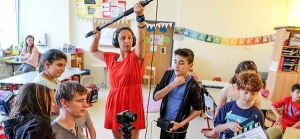 Das Foto wurde in einem Klassenzimmer aufgenommen. Eine Frau mit Kopfhörern hält eine Tonangel. Vor ihr steht eine Kamera. Kinder und ein Mann sitzen und stehen im Kreis um die Kamera herum. Ein Kind hält eine Filmklappe in der Hand.
