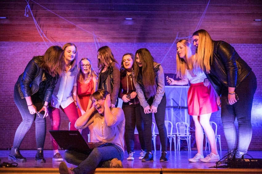 Referenzen-Theaterpädagogik-Prävention-Wo-ist-Hanna-03 Viele junge Frauen reden auf eine andere junge Frau ein. Diese sitzt mit einem Laptop am Rand einer Bühne und hält sich schreiend die Ohren zu.
