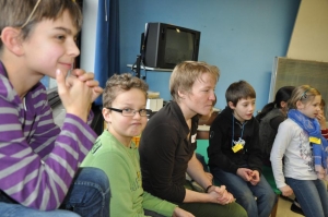 Kinder sitzen nebeneinander in einem Klassenzimmer. Zwischen ihnen sitzt eine Theaterpädagogin.