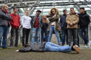 Jugendliche stehen im Kreis um eine Person, die am Boden liegt. Eine Frau mit verschränkten Armen blickt auf die Person herab.