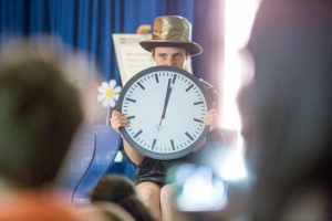 Eine Frau mit goldenem Zylinder hält eine große Uhr in der Hand.