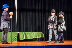 Kinder spielen gemeinsam ein Theaterstück. Zwei Kinder rechts im Bild tragen Sonnenbrillen, schwarze Hüte und gestreifte Jacken. Sie verschränken die Arme.