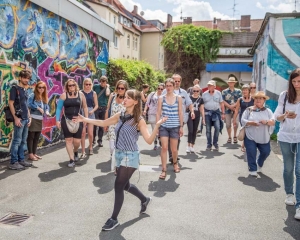 Viele Personen laufen gemeinsam durch eine Hildesheimer Gasse. Links befindet sich eine Wand mit Graffities. Im Hintergrund sieht man den Club "Thav". Die Personen werden von einer Frau angeführt, die mit ausladenden Gesten etwas erklärt.